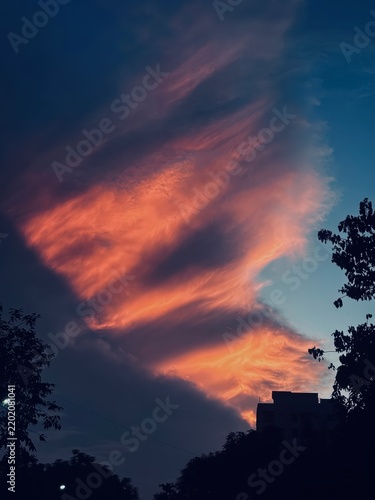 Dramatic vibrant sunset clouds painting colorful sky above silhouette of city buildings and greenery.