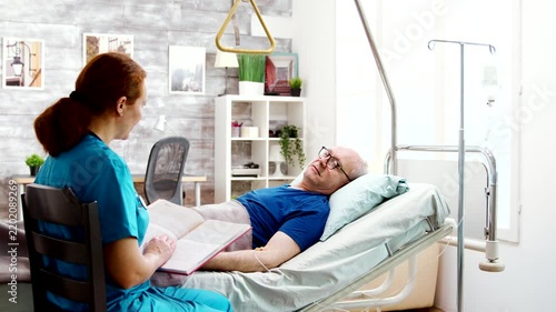 nurse reads a book beside an older man resting in an adjustable care bed, combining companionship, comfort and attentive senior healthcare.