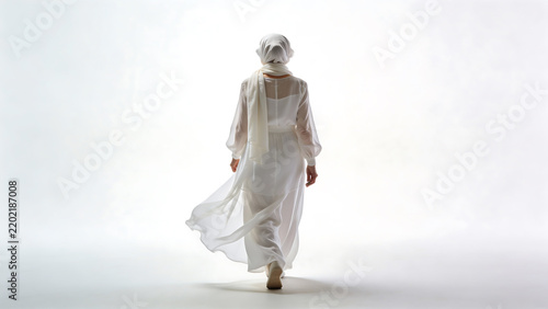 Woman in white hijab and flowing dress walking away against pure white background