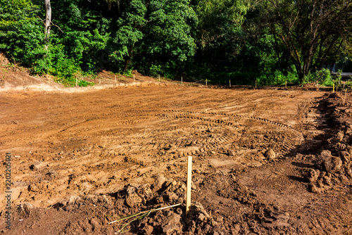 Freshly cleared construction site in a park featuring raw soil, deep tire tracks from heavy machinery, and survey stakes marking the perimeter for landscaping development
