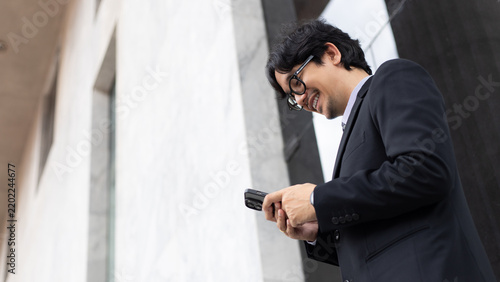 Smiling Asian businessman in a suit using a smartphone outdoors near a modern office building, checking messages, trading stocks, and managing mobile business communication online