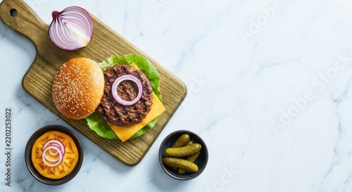 Top down view of a fresh homemade grilled beef burger with melting cheese, lettuce, sesame bun, red onion, and pickles on a wooden board against a white marble background with copy space.