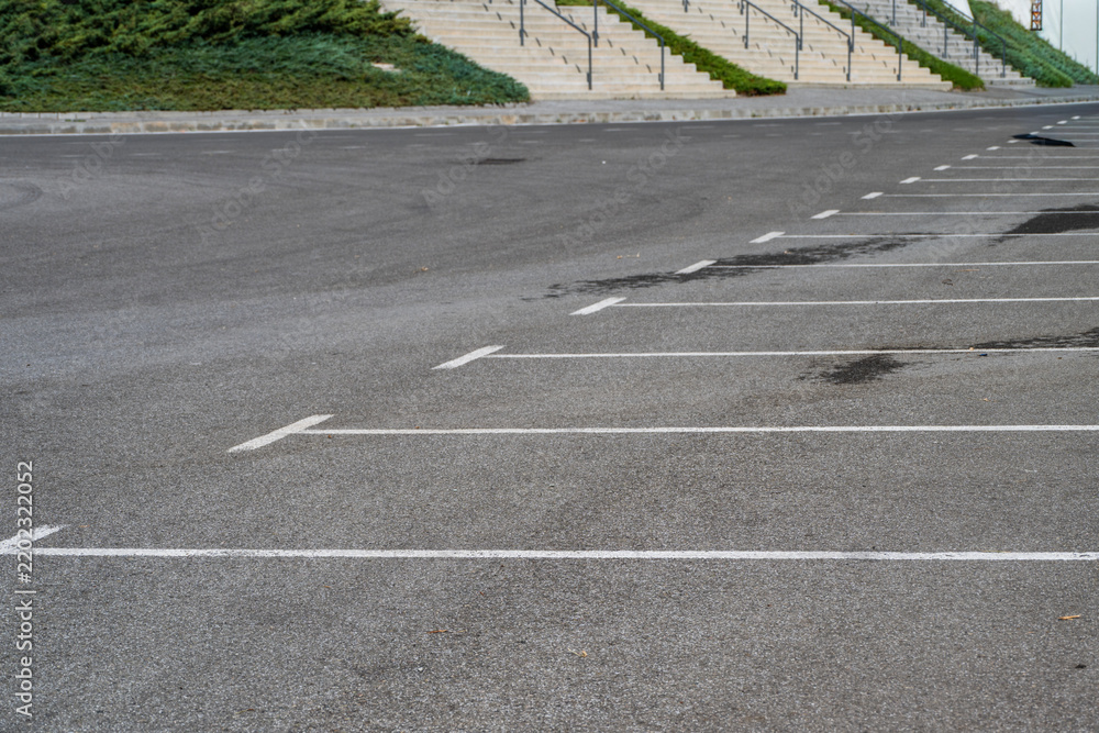 Obraz premium An empty outdoor asphalt parking lot is marked with white painted parking bays in front of landscaped hills and tiered concrete staircases