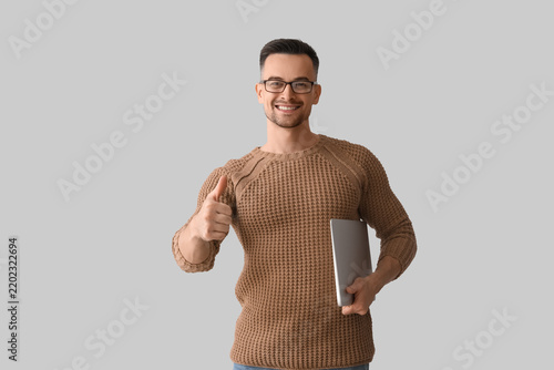 Happy young man with laptop showing thumb-up on light background