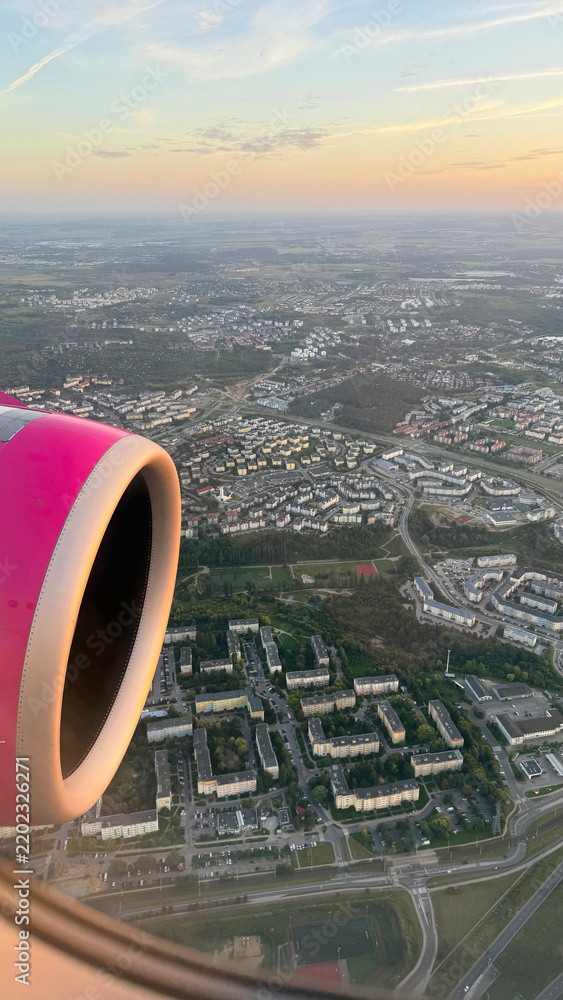 Obraz premium Aerial view of a city landscape with houses and streets through an airplane window next to a pink jet engine during flight, commercial aviation tourism and travel wanderlust concept