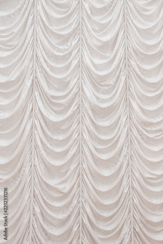 Cream fabric curtain features repeated scalloped swags and vertical pleats across the full surface, forming a soft ornamental backdrop with layered folds and neutral tones in an indoor hall
