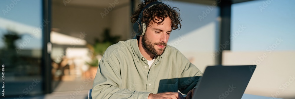 Fototapeta premium Wide panoramic remote work scene of man with headphones using laptop on a sunlit terrace for banner background and workspace design