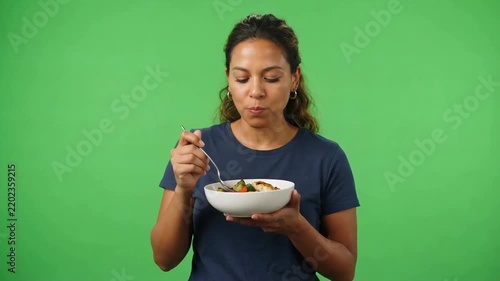Young woman eating a colorful salad from a white bowl against a bright green studio background, conveying healthy nutrition and enjoyment