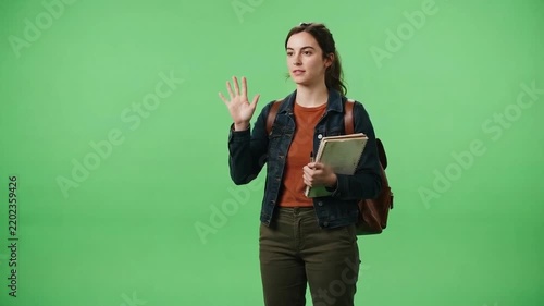 Young woman wearing a backpack, holding a notebook, and waving against a bright green screen background in a studio setting