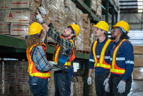 A diverse team of warehouse workers and engineers in safety helmets and reflective vests inspect palletized wood materials inside an industrial storage facility.