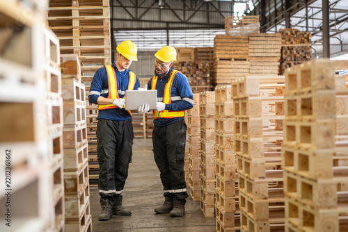 Two factory workers in safety helmets and high visibility vests inspect wooden pallets with a tablet in a large industrial warehouse.