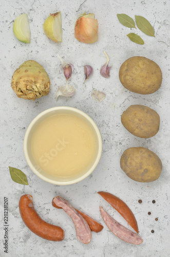 Top view ingredients for a regional dish known as *zalewajka* or *zurek*: rye sourdough starte, onion, garlic, potatoes, bay leaves, allspice and celery on grey background. It is a type of thick soup.