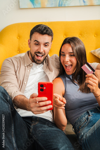 Two smiling partners lounge on a warm sofa, one holds a red smartphone while the other waves a card, creating a lively moment of shared activity and consumer interaction captured candidly.
