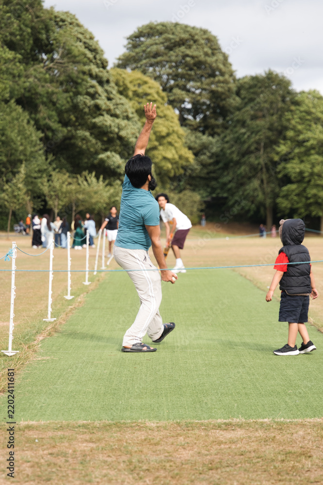 Obraz premium Adult man playing cricket, releasing the ball with a classic bowling action on a grassy pitch while a young child watches and other people gather in the background