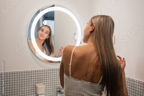Young woman standing in front of an illuminated bathroom mirror, holding and checking the ends of her long hair during a morning grooming routine.