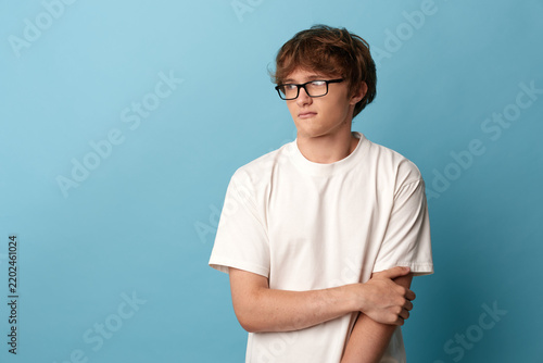 Bild auf Leinwand A pensive youth with glasses stands against a blue background, arms crossed, expressing contemplation, introspection, mood, and calm in a casual portrait