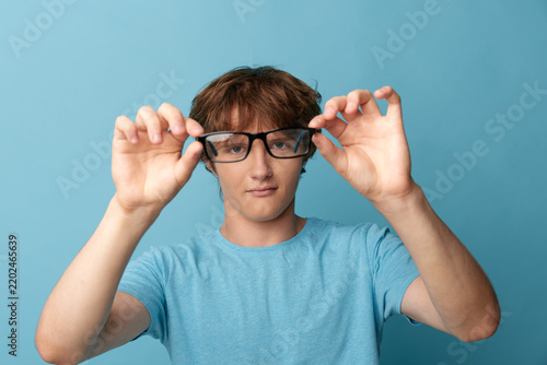 Bild auf Leinwand Young man holding black prescription glasses in front of his face, medium shot on blue background