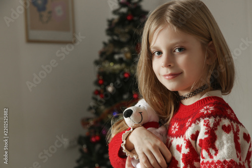 Side view profile of a cute blonde child sitting on a rug and gently holding a plush teddy bear with a Santa hat. Young girl wearing a red festive winter jumper with reindeers, cozy holiday atmosphere
