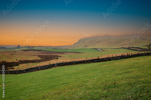 A view across the rolling green hills, dry stone walls and farmland of the Yorkshire Dales, England with a warm golden sky