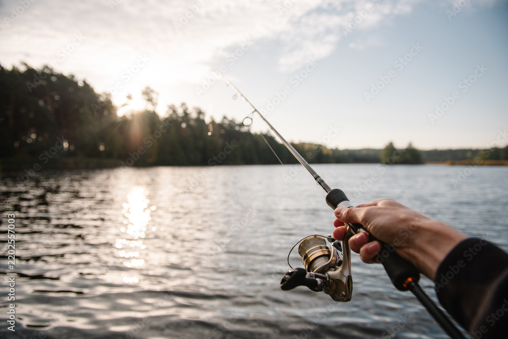 Fototapeta premium First person view of a fisherman holding a spinning rod with a reel over a serene lake at sunset, celebrating Fisherman's Day in nature.