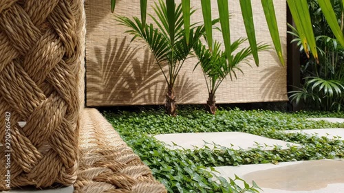 Palm fronds cast patterned shadows across a woven natural-fiber screen beside groundcover and pale stepping stones, shaping a tropical courtyard nook with tactile materials and restful greenery