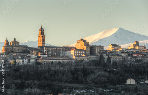 View of the historic town of Macerata with its bell tower and stone buildings illuminated by golden hour light, set against a backdrop of snow-capped mountains in Macerata, Marche, Italy.