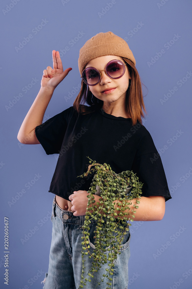 Obraz premium Young girl wearing sunglasses and a beige beanie poses playfully with a plant in her hand against a solid blue background