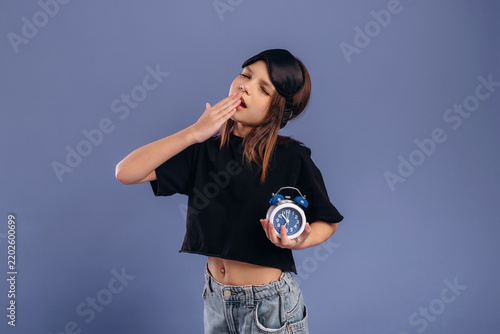 Young girl with long hair yawning while holding a blue alarm clock, wearing a black crop top and denim shorts against a solid purple background