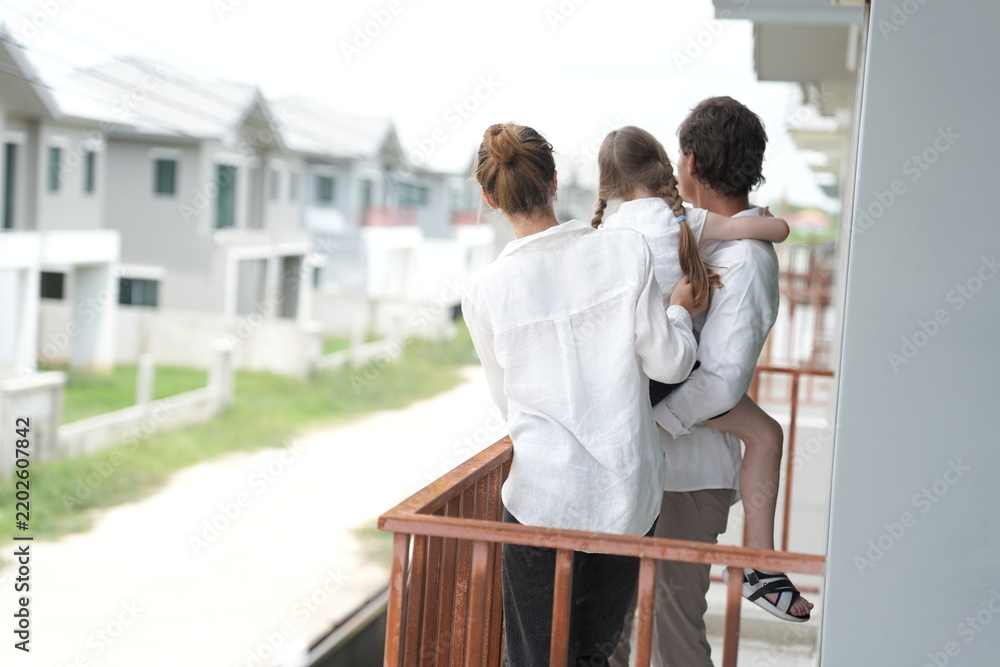 Obraz premium Happy young family with little daughter look out over the rows of new white houses from the railing on the upper balcony of their new home above the neighborhood, candid real moment