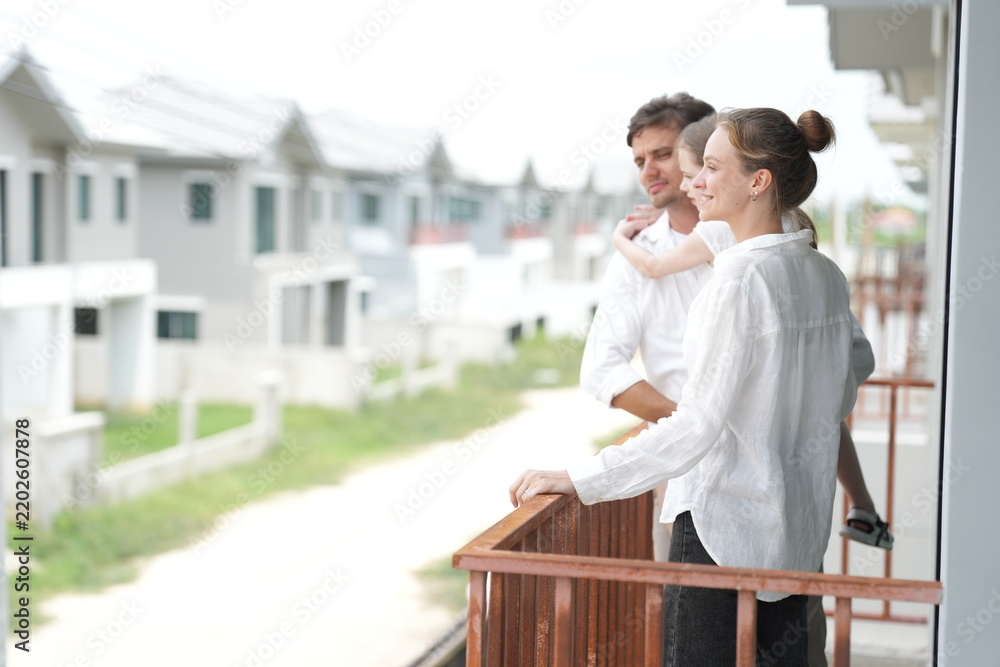 Obraz premium Young parents holding their child look out over the rows of new white houses from the railing on the upper balcony of their new home above the neighborhood, candid real moment