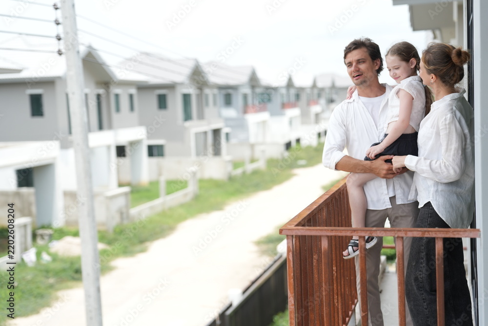 Obraz premium Caucasian family of three embracing enjoy the view of the growing suburb from their balcony standing on the terrace of a newly built suburban home in warm natural light