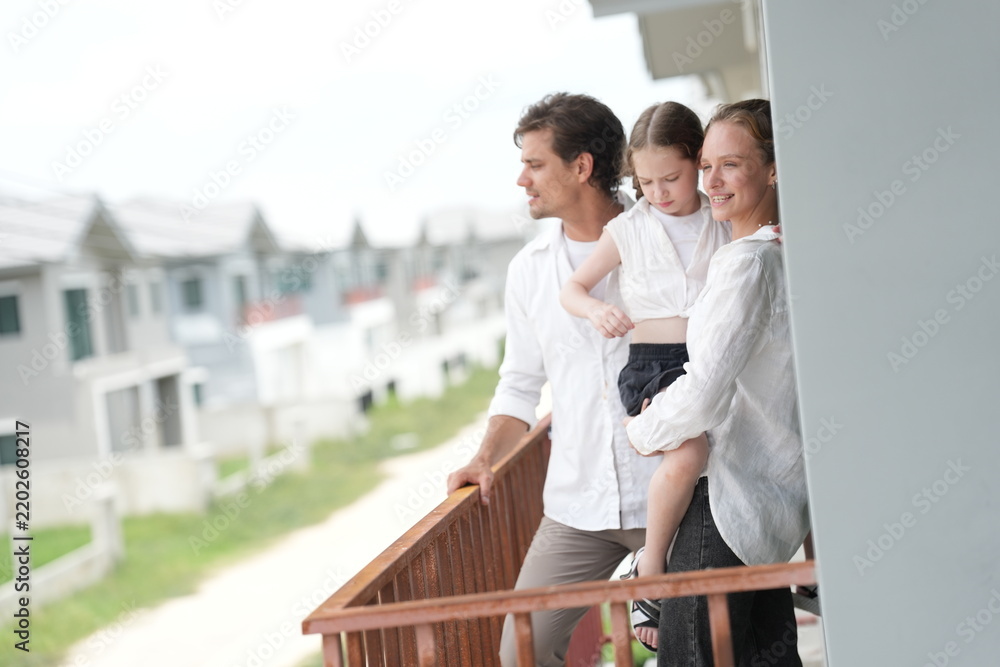 Obraz premium Caucasian family of three embracing stand at the wooden railing admiring the neighborhood view on the upper balcony of their new home above the neighborhood, authentic everyday scene