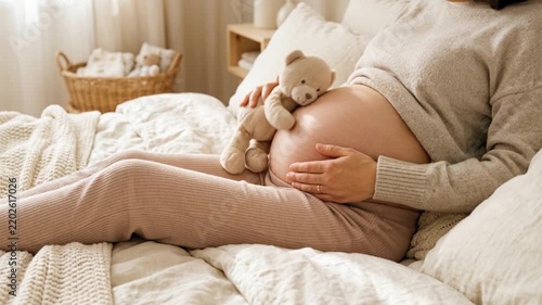 A woman in a gray sweater and beige pants sits on a bed holding her round belly while a teddy bear rests beside her, in a cozy room filled with soft light.