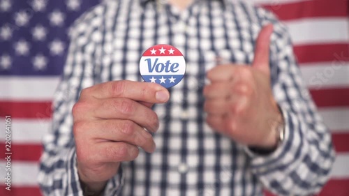 Man in plaid shirt holding Vote button badge in front of American flag