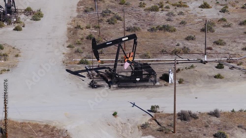 Aerial view captures an industrial oil pumpjack actively nodding on arid terrain beside dirt roads and utility poles in a sunny California oil field.