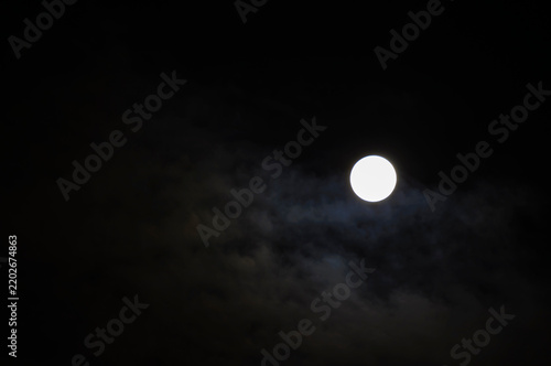 Midnight sky background. A bright white full moon glowing in a dark night sky. Thin clouds drifting across the sky around the bright lunar disc