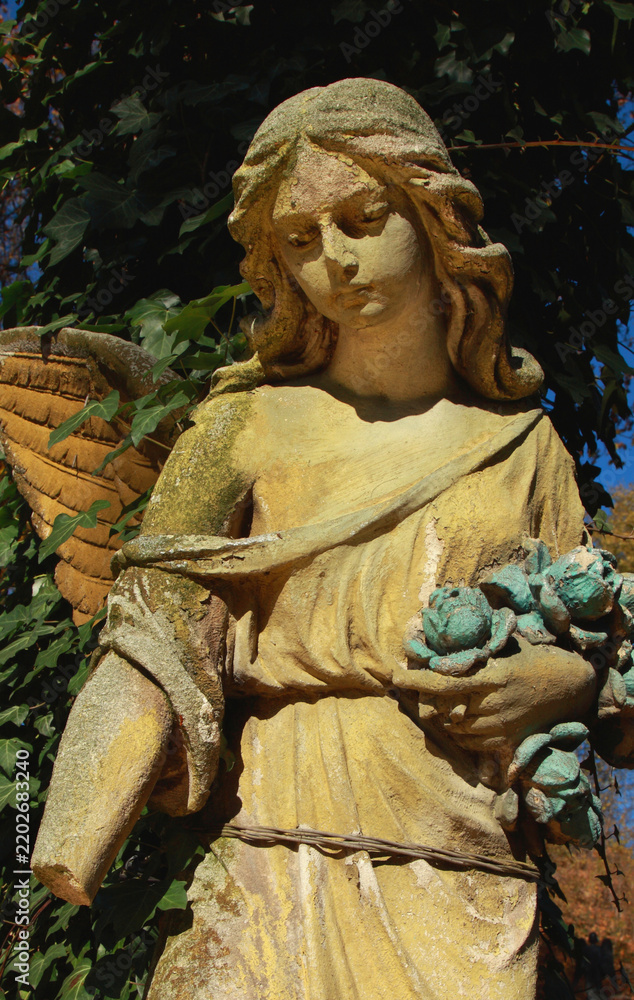 Obraz premium Weathered stone angel statue holding flowers, symbolic memorial sculpture representing remembrance, grief, eternal love, hope and peace.