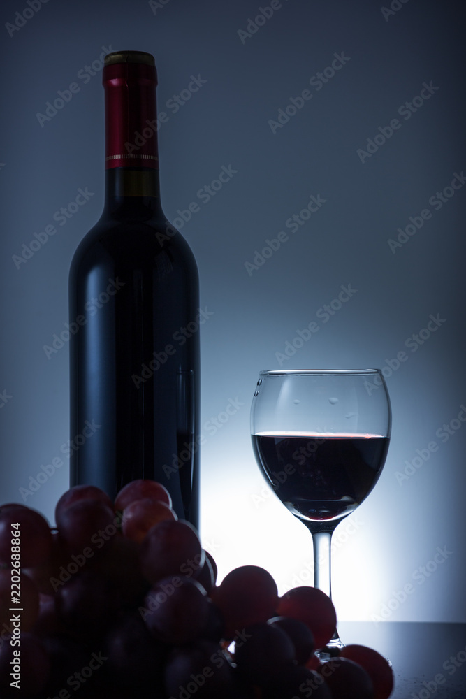 Fototapeta premium A still life composition featuring a red wine bottle, filled wine glass, and fresh grape cluster against a dark studio background.