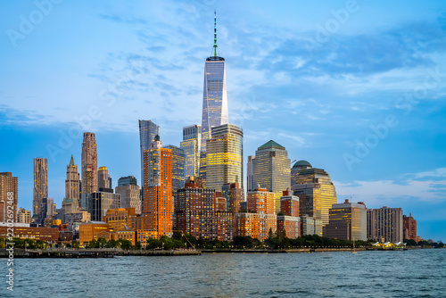 Office towers and waterfront apartments under an evening sky. Warm windows appear above brick waterfront blocks in New York City, with a glass spire against blue clouds.
