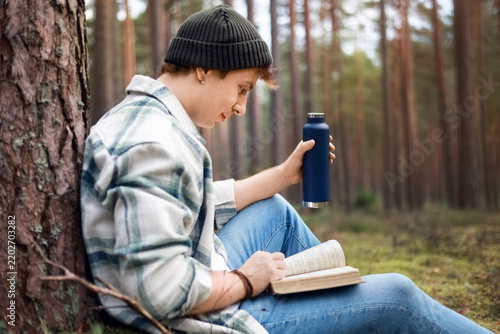 Man reading a book and drinking from a thermos in the forest, enjoying digital detox, mindfulness, nature therapy, stress relief, mental wellbeing, outdoor leisure, and relaxation.