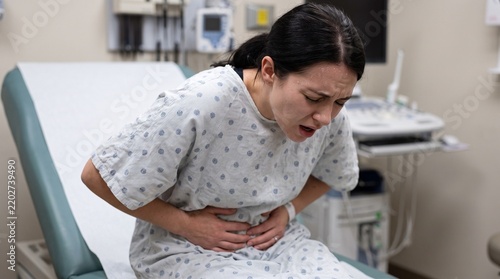 Young Woman in Hospital Gown Suffering Severe Abdominal Pain on Examination Bed