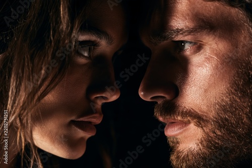 Close up portrait of woman and bearded man facing each other with intense eye contact and dramatic shadows against a dark background, intimate human connection