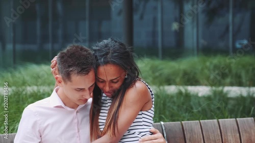 A mixed-race couple embraces affectionately while sitting on a wooden bench in a lush urban park during daytime. They share a tender moment surrounded by greenery and modern buildings.