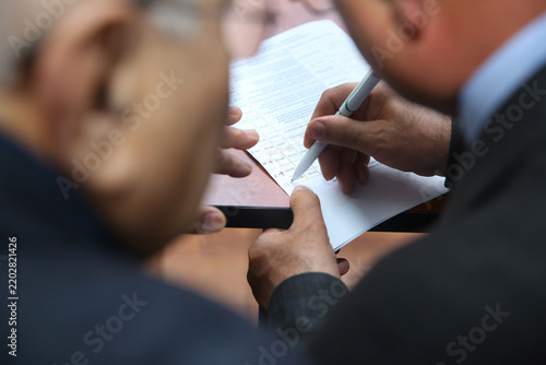 Two professional businessmen in suits work together closely. One man holds a pen to sign official paper documents and legal forms.