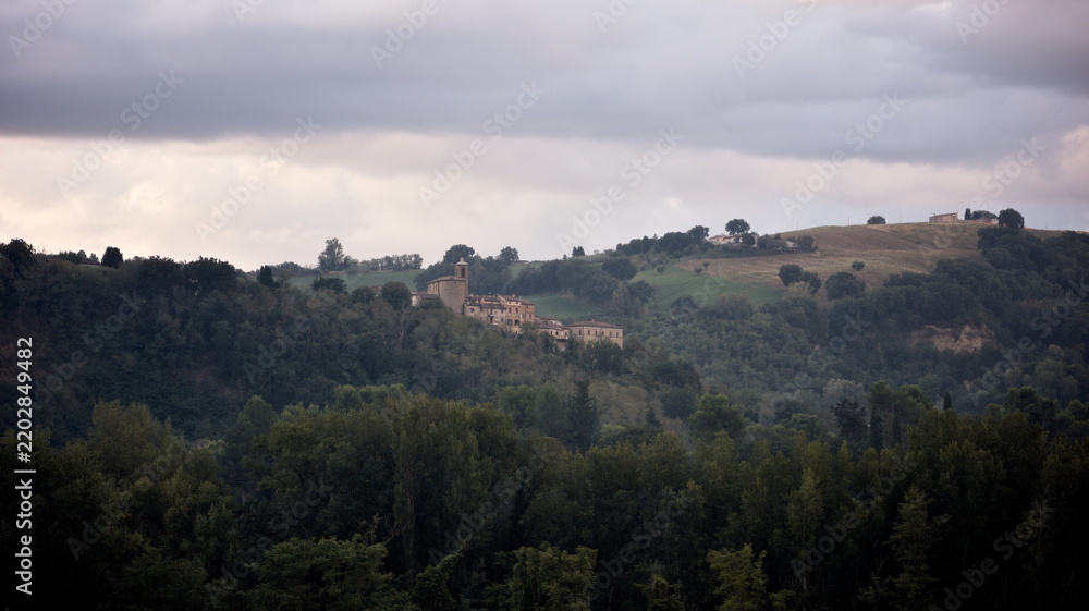 Fototapeta premium Paesaggio collinare italiano con piccolo borgo e case rurali sparse sui crinali, circondati da boschi lussureggianti sotto un cielo nuvoloso e drammatico
