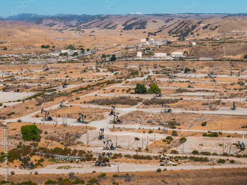 Aerial view shows numerous pump jacks extracting petroleum across an arid oil field landscape near rolling hills in California, alongside industrial storage tanks and infrastructure.