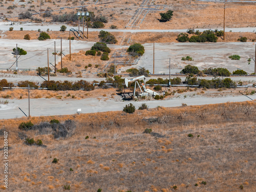 Aerial shot of an operating pump jack among pipelines and arid terrain in a Californian oil field, showing industrial petroleum extraction and energy infrastructure. Oil tariffs, trade war.