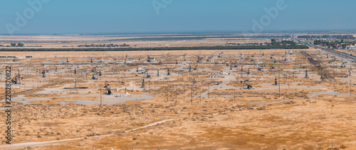 Aerial view of numerous pump jacks and extraction machinery situated across an arid oil field in California. Dirt paths cross dry flat terrain under a clear horizon during daylight.