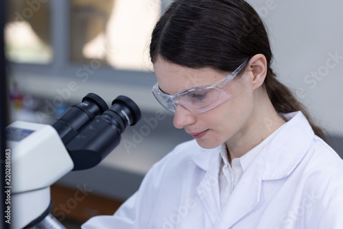 Quadro su tela Serious female scientist in white lab coat wearing safety goggles looking carefu