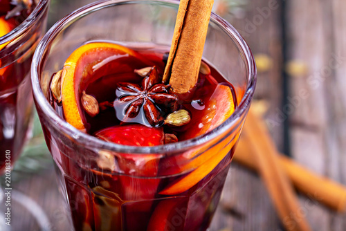 Close view of mulled wine in clear glass, garnished with orange slices, cinnamon, star anise, and cardamom, with additional cinnamon sticks resting on softly blurred wooden surface